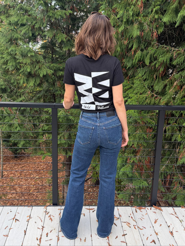 Person wearing a black tshirt with white text and blue jeans standing on a wooden deck with trees in the background.