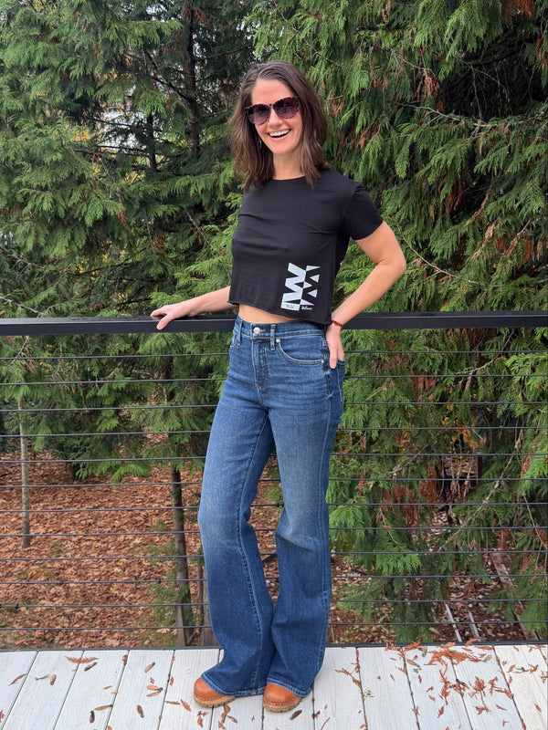 Person standing on a wooden deck wearing a black tshirt and blue jeans with trees in the background