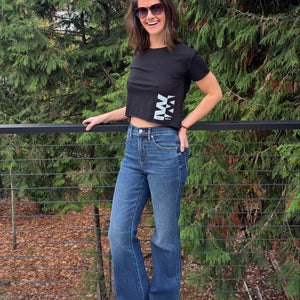 Person standing on a wooden deck wearing a black tshirt and blue jeans with trees in the background