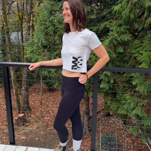 Person smiling and wearing a white tshirt and black leggings standing on a wooden deck with trees in the background