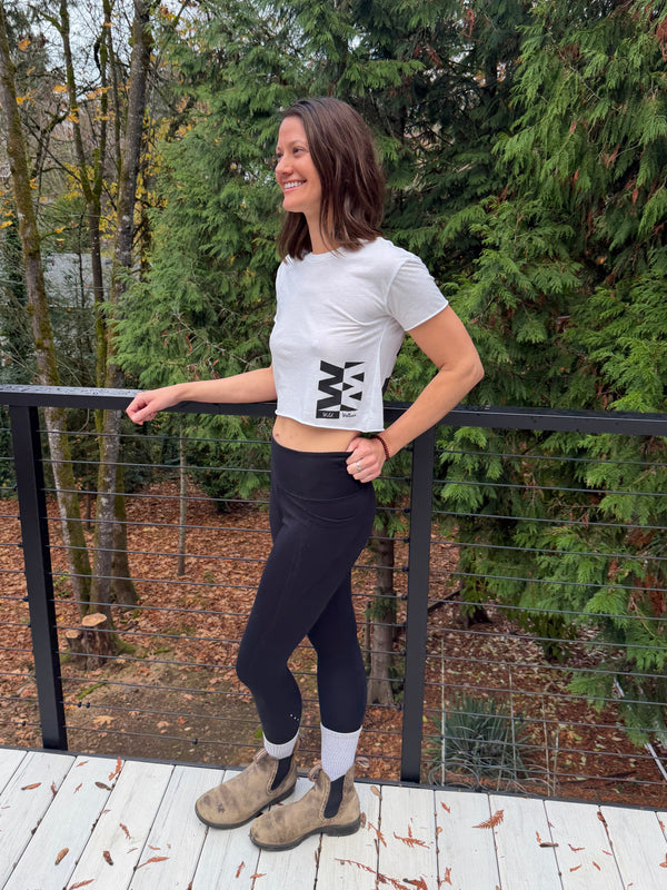 Person smiling and wearing a white tshirt and black leggings standing on a wooden deck with trees in the background