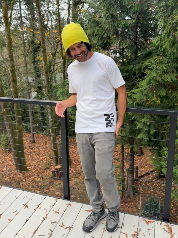 Person wearing a yellow beanie and white t-shirt standing on a wooden deck with trees in the background